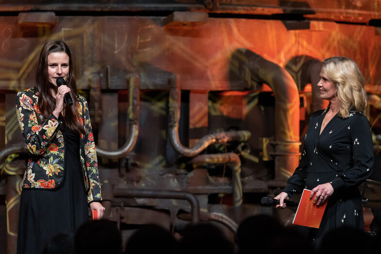 Photo of two female persons. One person is speaking into a microphone.