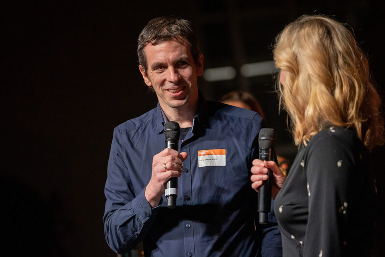 Photo of a male and a female person. Both people are speaking into a microphone.