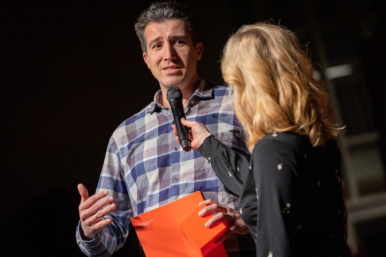 Photo of a male and a female person. The male person speaks into a microphone while holding a folder and a cube.