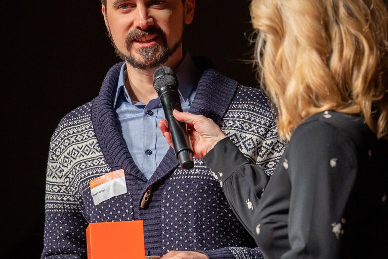 Photo of a male and a female person. The male person speaks into a microphone while holding a folder and a cube.