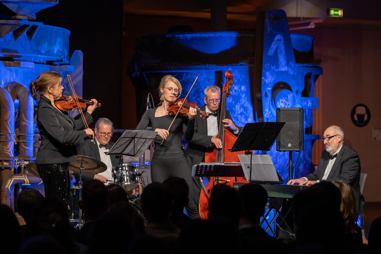 Photo of a band playing on a stage in front of an audience. The band consists of two females and three males. Each person plays a musical instrument.