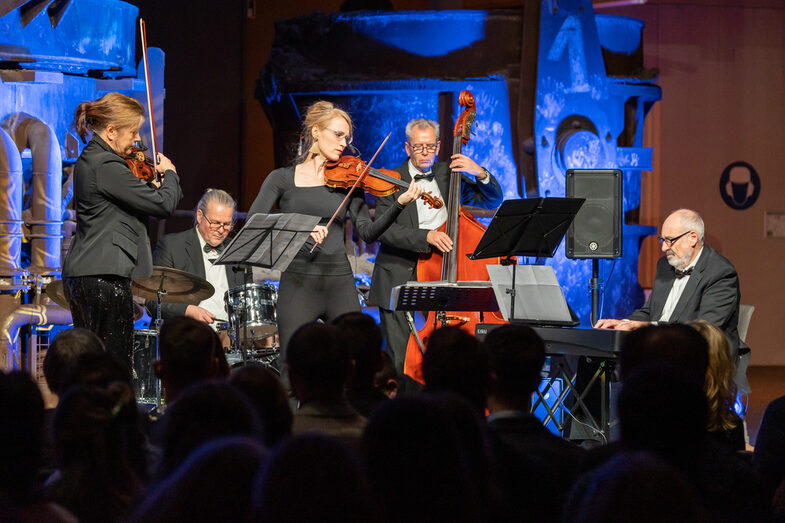 Photo of a band playing on a stage in front of an audience. The band consists of two females and three males. Each person plays a musical instrument.