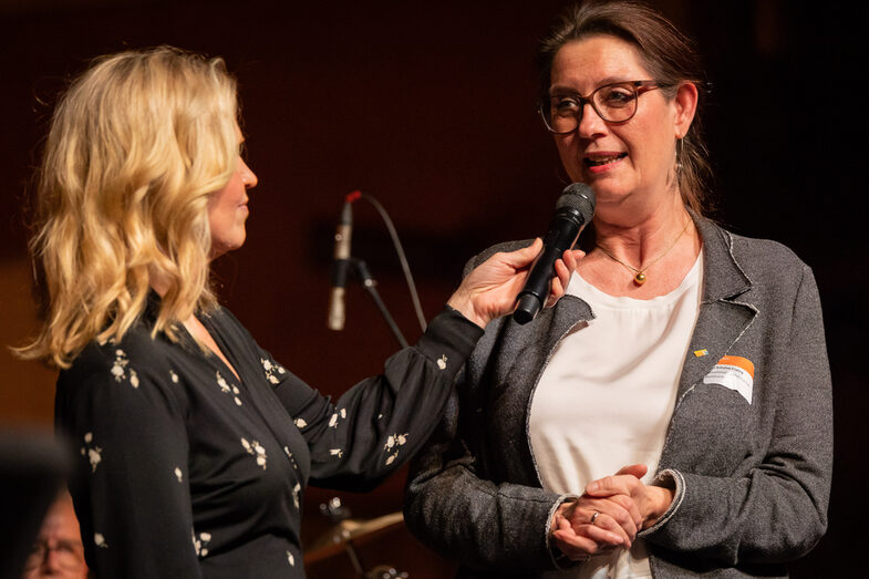 Photo of two female persons. One person is speaking into a microphone.