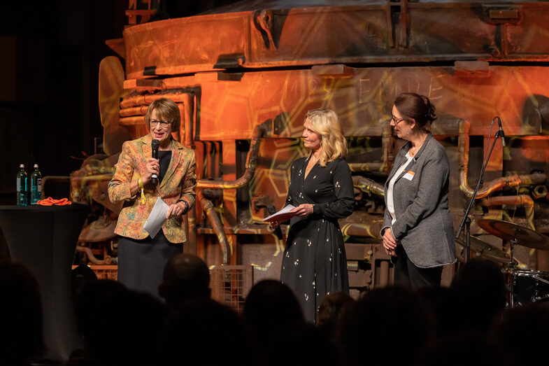 Photo of three female persons on a stage. One person is speaking into a microphone.