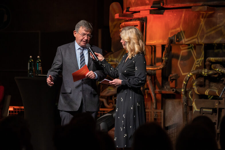 Photo of a male and a female person on a stage. The male person is speaking into a microphone.