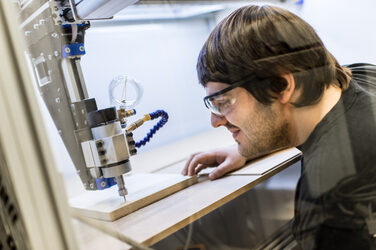 Foto eines Studenten, der ein Stück Holz in der CNC-Fräse fräst. Er trägt eine Schutzbrille.