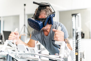 Foto eines Mitarbeiters mit VR-Brille. Er liegt auf dem Icaros-Fitnessgerät und hält sich an Handbefestigungen fest.