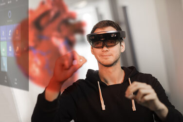 Foto eines Studierendem mit VR-Brille, der die Hände zur Steuerung in die Höhe hält. Vor ihm sieht man, was er durch die Brille sieht: ein Steuerelement der Software sowie ein 3D-Herz-Hollogramm. __<br>Student wears VR glasses and holds hands up to control. In front of him you can see what he sees through the glasses: a control element of the software and a 3D heart hollogram.