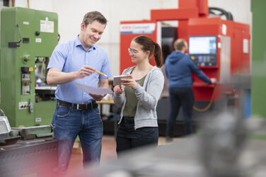 Photo of a man and a woman standing in a workshop. Both are holding pieces of paper. The man is pointing with a pen at the documents the young woman is holding. In the background, a man is standing at a large machine.