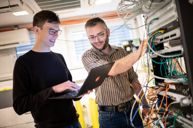 Foto von zwei Mitarbeitern im Serverraum, ein Mann steckt Kabel am Server, der andere überprüft am Laptop etwas.