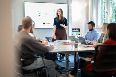 Eine Person steht vor einer Präsentation und erklärt etwas. Davor sitzen an einem Konferenztisch sechs zuhörende Personen mit Laptops und Notizblöcken