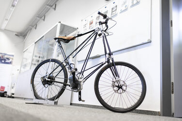 Ein Pedersen Fahrrad aus CFK und Aluminium mit einem elektrischen Antrieb über einen Mittelmotor steht vor einer Wand im Leichtbau-Technologie-Center. Im Hintergrund steht ein Regal und an der Wand befindet sich ein Whiteboard.