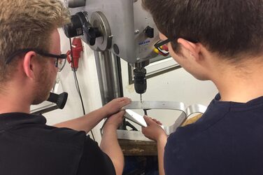 Zwei Studenten bohren Löcher in Teile ihres Geländers mithilfe einer Standbohrmaschine.