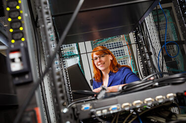 Foto einer Auszubildende im Serverraum am Laptop. Man sieht sie durch ein offenes Fach im Serverschrank. __ Trainee stands in the server room at the laptop. You can see them through an open compartment in the server cabinet.