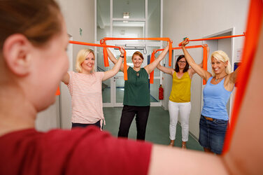 Foto von Mitarbeiterinnen, die auf dem Flur beim Pausenexpress mit orangenen Therabändern mitmachen. Sie halten die Bänder zum dehnen mit den Armen über den Kopf halten. __ Female employees take breaks in the hallway with orange Thera bands that they hold over their heads with their arms to stretch.