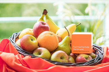 Foto von einem Korb, der mit Äpfeln, Bananen, Birnen und Orangen gefüllt ist. Im Korb liegt außerdem ein orangefarbener Würfel mit FH-Logo darauf. Der Korb steht auf einem orangenen Tuch.