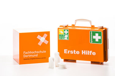 Foto eines orangefarbenen Erste-Hilfe-Koffers, Verbandszeug und eines orangefarbenen Würfels mit FH-Logo. Auf dem Würfel kleben zwei Pflaster überkreuz.