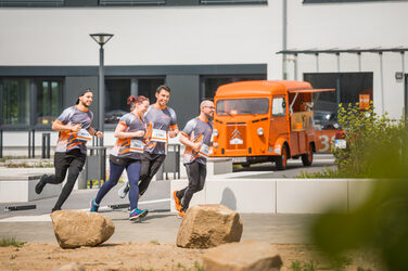 Foto von vier Mitarbeiter*innen in FH-Laufkleidung, die bei einem organisierten Lauf über den Campus mitlaufen. Im Hintergrund sieht man das Gebäude 38 b und den orangenen Oldtimer.. __ <br>Employees with FH equipment run along in an organized run across the campus. In the background you can see building 38 b and the orange classic car.