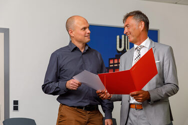 Foto des Kanzlers der Fachhochschule Dortmund, der ein Zertifikat an einen Mitarbeiter übergibt, der  neben ihm steht. In der anderen Hand  hält der Kanzler eine orangene Mappe. __ The Chancellor - on the right in the picture - hands over a certificate to an employee who stands to his left and accepts it with his right hand. The Chancellor is holding an orange folder in the other hand.