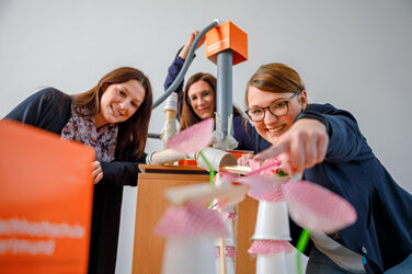 Foto von Kolleginnen, die beim Teamtag eine bunte Murmelbahn aus unterschiedlichen Materialien gebaut haben und nun erfreut damit spielen. __<br>At the team day, colleagues built a colorful marble run from different materials and are now enjoying playing with it.