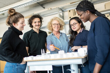 Foto einer Professorin, die gemeinsam mit ihren Studierenden hinter einem Architekturmodell steht und mit einem Stift auf einen Part zeigt, um etwas zu erklären. __ A professor, surrounded by her students, stands behind an architectural model and points with a pen at a part to explain something to her students.