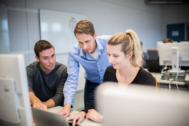 Photo of a professor standing between a male student and a female student in the computer room and explains something to both of them on the laptop. __ <br>The professor stands between a male student and a female student in the computer room and explains something to both of them on the laptop.