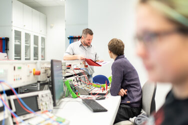 Foto eines Professors, der in seinem Laborraum an einer Aufbauwand lehnt, er hat eine Mappe in der Hand. Ein Studierender, der vor ihm am Tisch sitzt, schaut zu ihm auf. Auf der langen Tischreihe stehen diverse elektronische Versuchsaufbauten. Vorne im rechten Bildrand, unscharf erkennbar, ist noch eine Studentin. __ <br>A professor is leaning against a wall in his laboratory with a folder in hand. A student sitting at the table in front of him looks up at him. There are various electronic test setups on the long row of tables. In the front right of the picture, out of focus, is another student.