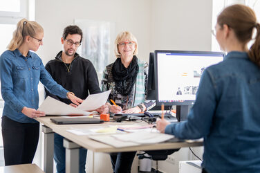 Foto von vier Mitarbeitenden in der Verwaltung am Schreibtisch, eine Person davon in Rückansicht. Diese unterhält sich mit einer Frau ihr gegenüber, die anderen beiden Mitarbeitenden sichten Unterlagen.