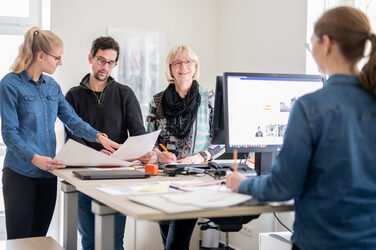 Photo of four employees in the administration department at their desks, one of them seen from behind. This person is talking to a woman opposite her, the other two employees are looking through documents.