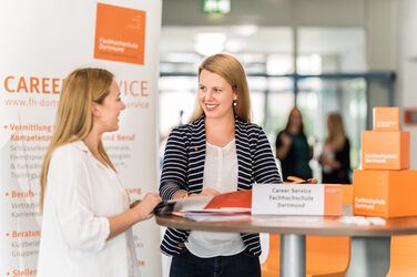 Photo of an employee of the Career Services of the Fachhochschule Dortmund standing at a high table talking to a young woman. __ <br>Employee of the Career Services of the University of Applied Sciences Dortmund stands at a high table talking to a young woman.