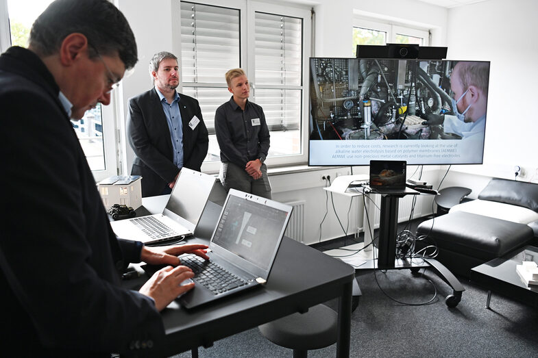 Auf einem großen Bildschirm läuft ein Video während daneben ein paar Menschen zusehen sind. Ein weiterer Arbeitet im Vordergrund an einem Laptop.