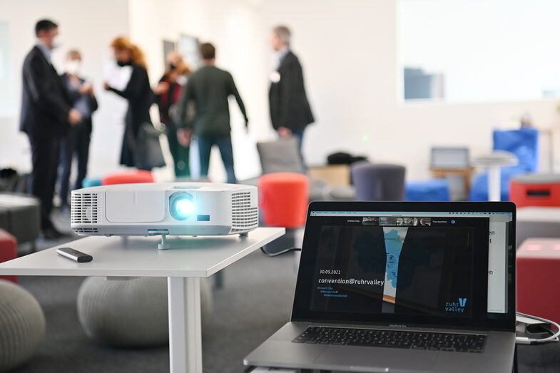 Ein Laptop steht neben einem Beamer. Auf dem Monitor ist das ruhrvalley Convention Logo zu sehen. Hinter Laptop und Beamer steht eine Gruppe Menschen und unterhält sich.