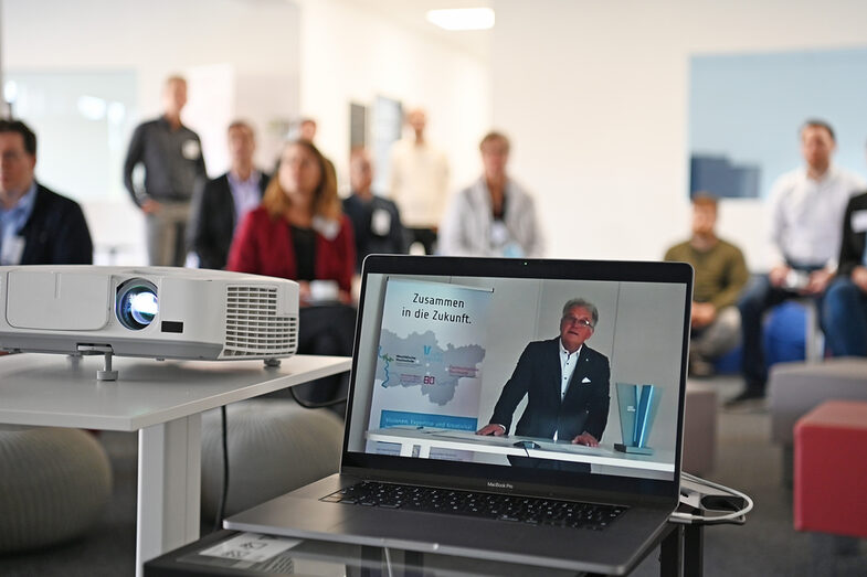 Ein Laptop steht neben einem Beamer. Auf dem Monitor ist ein Mann zu sehen, der spricht. Hinter Laptop und Beamer sitzen Menschen die nur unscharf zu erkennen sind und hören zu.