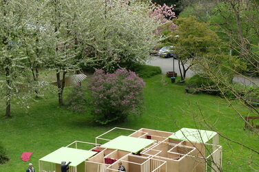 Der Irrgarten hat seine Form gefunden. Studierende bringen gemeinsam die letzten Einzelteile an. Neben den Holzwänden und Lattenrahmen sind weitere Bausteine hinzugekommen. In der Mitte des Entwurfs sind Vorhänge zu erkennen, außerdem fungieren Lamellen als Trennwände und grüne rechteckige Flächen bilden eine Dachebene. Platzierte Leerflächen innerhalb des Entwurfs sorgen für eine offene Wirkung und verstärken die Illusion, dass der Raum im Zentrum zu erreichen sein könnte.