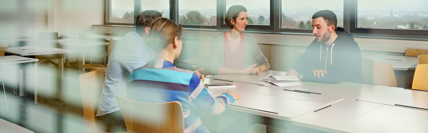 Photo through a window pane into a seminar room where four students are sitting at a table and exchanging ideas.