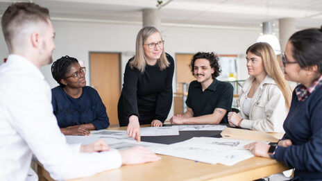 Foto von einer Mitarbeiterin des Fachbereichs Architektur, die eine Mappenberatung mit mehreren Studierenden an einem Tisch durchführt. Sie zeigt auf eine Mappe, die auf dem Tisch liegt.