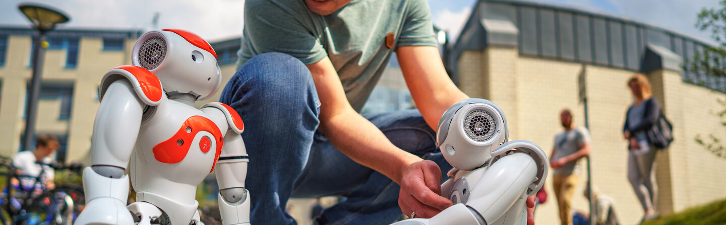 Photo of two NAO robots standing outside Building 42. An employee is setting up the robots. A Fachhochschule Dortmund cube is placed between the robots.