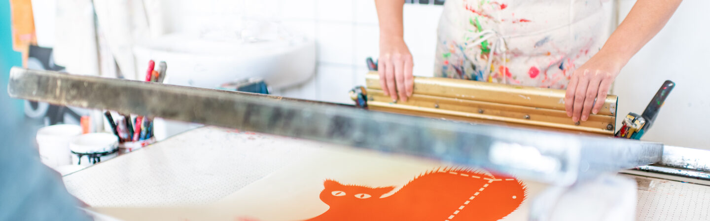 Photo of two people creating a silkscreen of an orange cat. Only the upper body and arms of the people are visible.