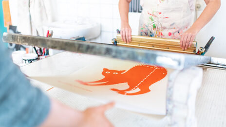 Photo of two people creating a silkscreen of an orange cat. Only the upper body and arms of the people are visible.