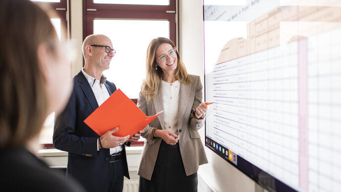 Photo of employees. They are talking smiling - the man has a folder in his hand, the woman right next to him is pointing at the large monitor. Across from them, another woman is strongly cropped.