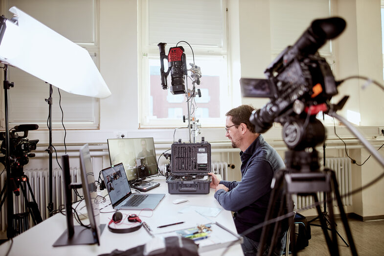 A prof sits at his desk, surrounded by various camera equipment as well as a laptop and monitors, in order to give an online lecture.