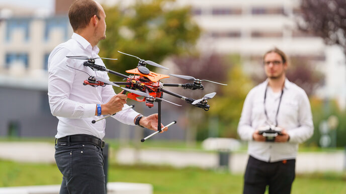 Photo, two males. The person on the right holds a flying drone.