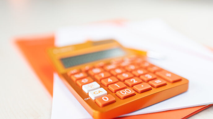 Foto eines orangefarbenen Taschenrechners, der auf einer orangefarbenen Mappe und einem Stapel Zettel liegt. __ <br>An orange calculator lies on top of an orange folder and a stack of papers.