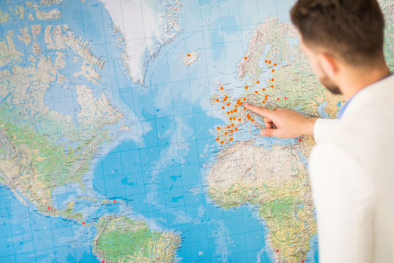 Foto einer Person, die auf eine große Weltkarte zeigt, auf der mit orangefarbenen Punkten Orte markiert sind.