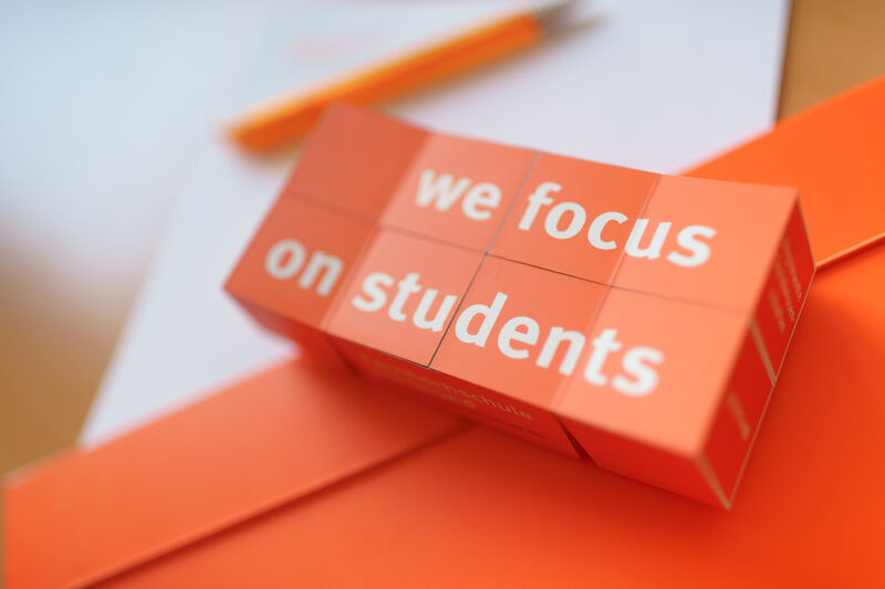 Foto eines aufgeklappten Klappwürfels mit dem Aufdruck "we focus on students", der auf Unterlagen liegt. __ Opened folding cube with the imprint "we focus on students" lies on documents.