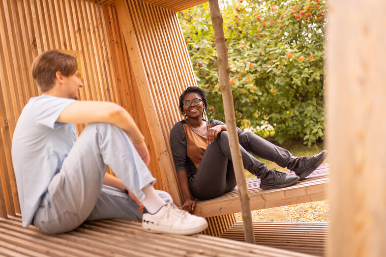 Foto von zwei Studierenden, die in einem Holzgebilde sitzen und sich locker unterhalten.