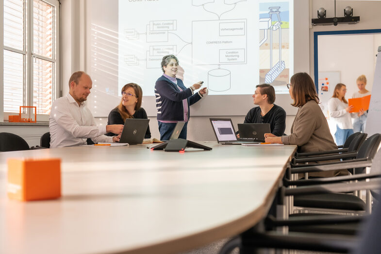 Foto einer Besprechungssituation mit 5 Personen. 4 sitzen an Laptops und eine Frau präsentiert im Stehen. 2 Frauen sind im Hintergrund in einem weiteren Raum erkennbar.
