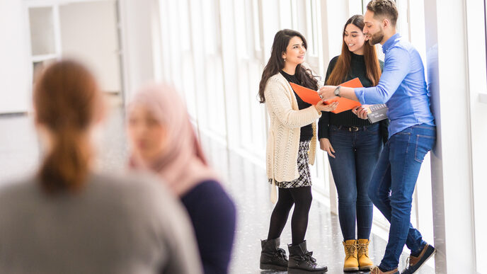 Photo of five international students, two of whom are talking in the foreground and the other three are looking together into an orange folder.