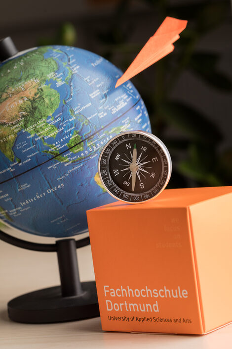 Photo of a still life: globe in the background, Fachhochschule Dortmund cube in front, orange paper airplane and compass.