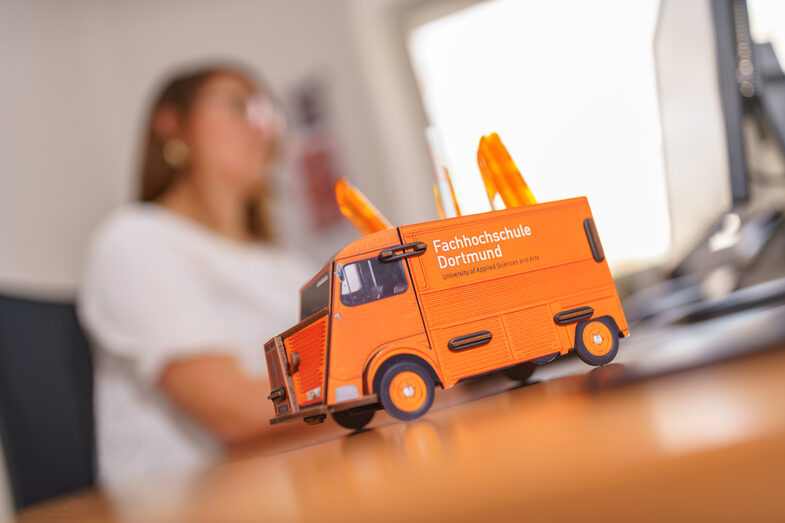 Photo of a pen box in the form of a Citroen HY panel van on a desk, in the background a woman is working on the computer.